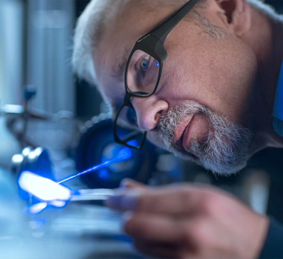 A man in glasses aligns a laser on a piece of glass in a lab, wearing protective glasses.
