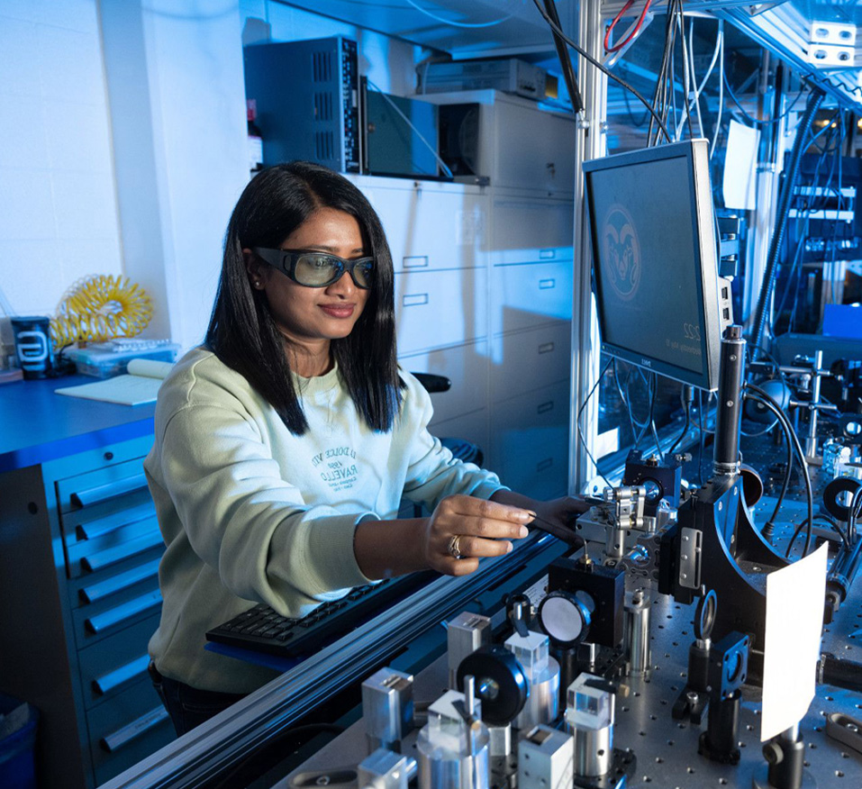 A woman in a lab adjusts a laser while working on a computer and operating a machine.